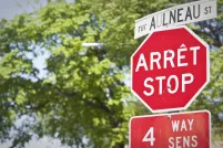 A stop sign and street sign in both English and French are seen in front of trees.