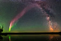 A vivid arc of pinkish-red and green aurora curves across a starry night sky alongside the Milky Way's core, above a dark tree-lined lake that mirrors the lights on its calm surface.