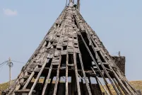 A large, weathered wooden structure shaped like a tepee stands in a dry grassy field under a clear blue sky. The structure's wooden frame is largely exposed, with many wood shingles missing or falling off, revealing the ribbed skeleton beneath.