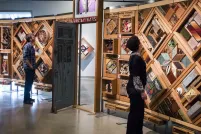 Three people in a gallery examining the Witness Blanket, a large free-standing blanket made of cedar panels holding objects related to the residential school experience.