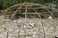 A dome frame of lashed wooden poles over a central pile of stones, surrounded by trees with water beyond.