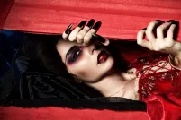 A vampire dressed in a velvety red dress peeks through a red wooden box with her long, black nails.