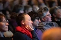 Audience members sit in a dimly lit venue watching a presentation, their faces illuminated by stage light. In the foreground, a person with their hair in a bun and wearing a bright red scarf rests their chin on their hand, looking attentively toward the stage. Other attendees sit beside them in soft focus, creating a quiet, contemplative atmosphere as the crowd listens.