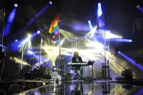 A performer is sitting behind a keyboard on stage with beams of blue and white lights and a rainbow flag flying on a pole.