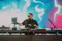A bearded, dark-haired man, wearing sunglasses and a black t-shirt, is performing on stage using a computer and an electronic keyboard mixer.