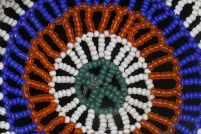 A close-up photo of a circular beadwork design on a black background. The pattern features concentric rings of turquoise, white, orange and bright blue beads arranged in a radiating floral or sunburst motif.