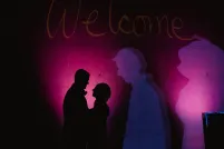 A man and woman in silhouette face each other in front of a dark wall illuminated by purple light.