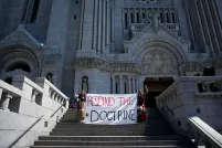 Two people in braids and ribbon skirts raise fists and hold a large cloth banner reading “RESCIND THE DOCTRINE” on the steps of an enormous cathedral.