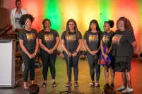 A group of six black performers standing on stage with colourful backlighting.