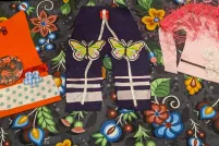 Pieces of crafted Indigenous- style clothing, including an orange shirt, pair of pants and made from colourful fabric and ribbon is are laid against a piece of craft paper adorned with flowers and berries. The clothing is includes an orange shirt, a pair of pants with ribbons and a pink tunic decorated with colourful fabric, appliqués and ribbons. The tunic has with a feathered shawl on the shoulders.