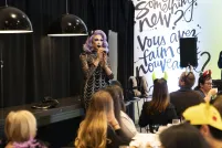 A drag performer with purple hair and a silver and black dress speaks in front of a group having brunch.