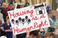A group of people walking down a street. One person holds a sign that reads: “Housing is a human right.”