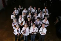 A group of twenty-two people facing the camera and smiling. They are all wearing dark-coloured pants and white shirts with red embroidery. A majority of the group are holding their instruments, mainly mandolins. 
