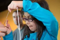 A child making a craft works intently to pull a string. The child is dressed in a blue sweatshirt with a snowman on it.