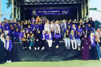 Dozens of people are gathered on a stage, most smiling and wearing the colour purple. Many are wearing hijabs. A banner hangs at the back of the stage that reads "Take Initiative. End Islamophobia. #OurLondonFamily."