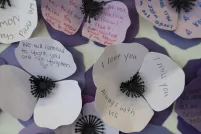 Three-petal flowers made of paper in various shades of purple are grouped together in a wall display. Names and messages are written on them. 