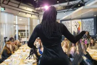 A drag artist with long locs in a black long-sleeve outfit faces away from the camera towards a seated audience at a banquet-style event, with a colourful raised-fist mural visible on the far wall and a stage light glowing overhead.