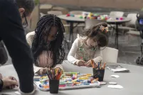 Two young children sit side by side at a round table, engaged in an art activity with markers and coloured pencils.