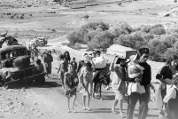 A black-and-white photo of a group of adults and children carrying belongings, walking down an unpaved desert road past a broken-down truck.