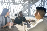 Three people are seated at a table. They're all smiling with coffees in front of them. There is a small progress pride flag on their table and at the empty table behind them. The sun is shining bright through the Museum windows.