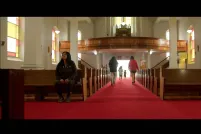 A church interior with a long red-carpeted aisle between wooden pews. Sunlight filters through stained-glass windows as several children run toward the bright entrance at the far end while an Indigenous woman sits alone in a pew to the left.