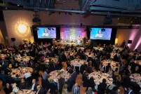 In a large hall, hundreds of diners sit at round tables while a woman speaks at a podium.