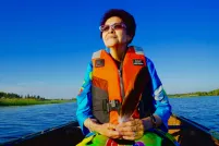 An indigenous woman is in a canoe on a lake with an orange life jacket. She seems at serene.