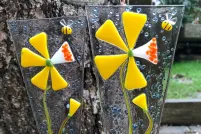Two fused glass ornaments depicting yellow flowers sit in a flower pot outside.