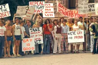 A large group of people dressed in T-shirts and seventies-style flared pants, stand together holding signs with messages such as “Out of the Closet,” “Gay is Good” and “Repeal Anti-Gay Laws.”