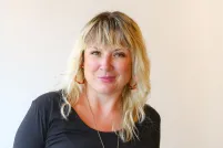 A smiling blonde woman wears a comfortable and elegant black scoop-neck top and a long brass necklace with a large white stone.