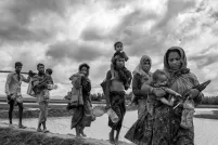 A group of Rohingya women and men carry young children and belongings as they walk in a line over an earthen dike over a stretch of water.
