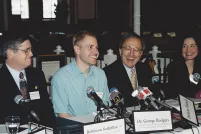 A man wearing a light blue shirt sits at a long table in front of a cluster of microphones, smiling. Seated on both sides of him are other people dressed in suits and facing microphones, also smiling. The name placards along the front of the table read, from left to right, “Murray Billet”, “Robinson Koilpillai”, and “Dr George Rodgers”, with too-blurry-to-read affiliations below the names. Another placard, knocked on its side, reads “Delwin Vriend” without an affiliation.