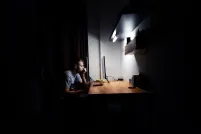 A white man with short hair sits alone at a brightly lit desk in a dark room, staring at a computer monitor.