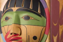 A closeup of a carved wooden box, showing the carved face of a person with a painted red hand over their mouth.