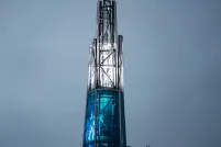 A tall, illuminated glass-and-steel spire rises above the curved patterned roof of the Canadian Museum for Human Rights, glowing in blue and white light against a soft grey sky.