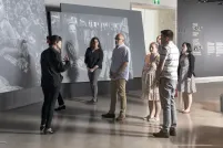 A small group of people stand in front of large screens displaying photographs and look at a woman who is speaking to them.