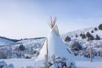 A snow-covered tipi sits in a hilly landscape dotted with trees and bushes against a blue sky.