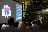 Audience members view a large projection screen displaying an elderly woman in a purple top and blue pants sitting in a white armchair, alongside an interactive message interface. The presentation takes place in a modern space with geometric glass architecture, and a presenter stands to the right of the screen.