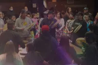 A group of men drum and sing in a circle. They are tightly surrounded by a large and diverse crowd of people all singing along.