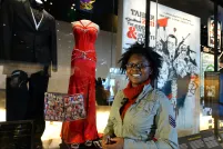 A smiling woman stands in front of a red prom dress and a black tuxedo mounted on mannequins. Both the dress and suit are on display behind a glass case.