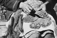A black-and-white photo of hands holding and stitching decorated fabric pieces with floral embroidery and textured fibres spread across a person's lap.