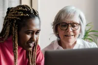 A young woman who looks to be in her teen years smiles as she works at a computer alongside an older, white-haired woman who is also smiling.