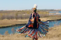 A Indigenous dancer in brightly coloured Fancy Shawl regalia beside a river.