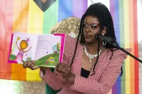 A drag queen in heavy makeup and a wig of long tiny braids holds up a children’s book to show an audience. The backdrop behind her is a pastel rainbow.