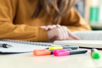 Close-up of a spiral notebook, a pen, and colorful highlighters on a desk, with a person in a mustard sweater sitting blurred in the background.
