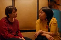 Two Inuit women sit in a dim room facing one another in intimate conversation. One is wearing a red sweater, the other a yellow sweater.