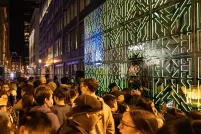 Many people stand between two intricately patterned joined mirrors that reflect green lights and a throng of onlookers.