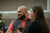 A man and woman sit together at a dinner table, smiling warmly as they listen to other guests. The man wears a salmon shirt with a gray vest, and there are small candles and drinks on the table, creating a cozy intimate atmosphere.