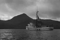 A boat in the water with mountains and clouds in the background. Its sail is decorated with large peace signs.