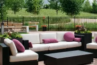Couches and chairs sit in a fenced patio. Small trees and tall green prairie grass surrounds the patio. In the background, the spire from Esplanade Riel rises above the trees.